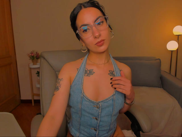SarahLuna with glasses, wearing a blue denim halter top, showing small tits with butterfly tattoos on her chest, black nail polish, and hoop earrings, sitting in a chair.