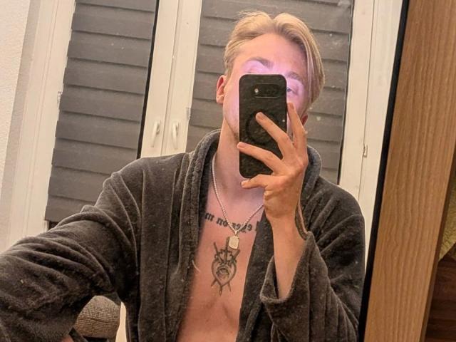 Honig96-hot with blonde hair, wearing a grey robe, chest tattoo, and necklace, taking a mirror selfie with a phone, showing a clean-shaved face and purple light reflection.