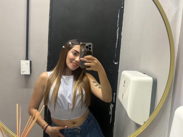 SharonKlart with long brown hair, white crop top, and blue jeans, smiling in mirror selfie, showing tattoo on left arm, hand in jeans, holding phone, in bathroom.