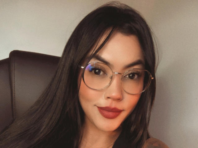 EmilyLo, con lunghi capelli neri, che indossa occhiali, rossetto rosso e un sorriso discreto, seduta su una poltrona in pelle marrone. Il suo seno non è visibile..
