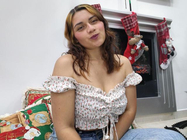 Ehmily, com seios médios, vestindo uma blusa branca com estampas florais e alças que caem do ombro, jeans azuis, cabelo castanho ondulado, batom rosa claro, sentada no sofá, com meias de Natal, e uma lareira ao fundo..