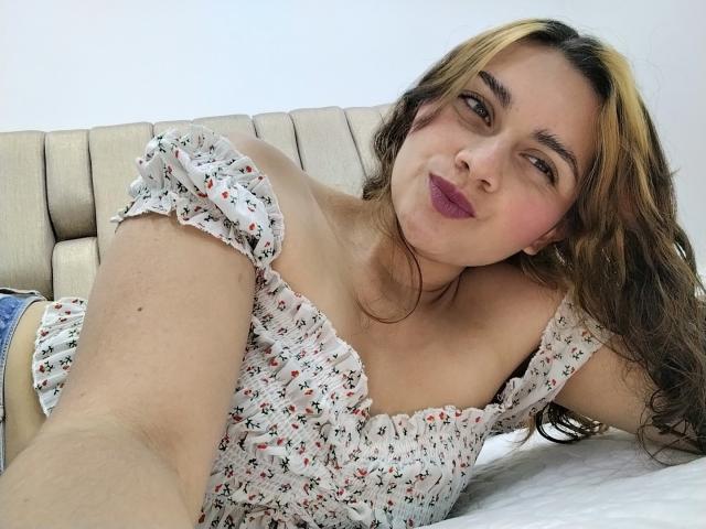 Ehmily, com cabelos castanhos longos, lábios roxos finos e seios pequenos, vestindo uma blusa floral com um ombro de fora, deitada em uma cama, tirando uma selfie, olhando para a câmera..