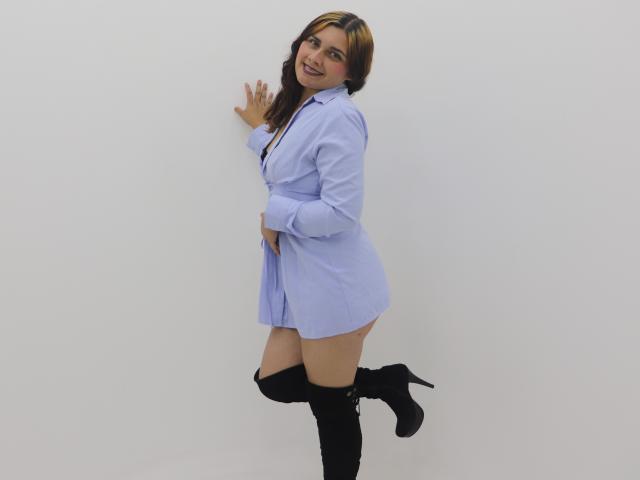 Ehmily tiene el cabello castaño claro con reflejos rubios, lleva una camisa azul desabrochada, botas negras que le llegan al muslo y no lleva pantalones, está de pie frente a una pared blanca y está sonriendo..
