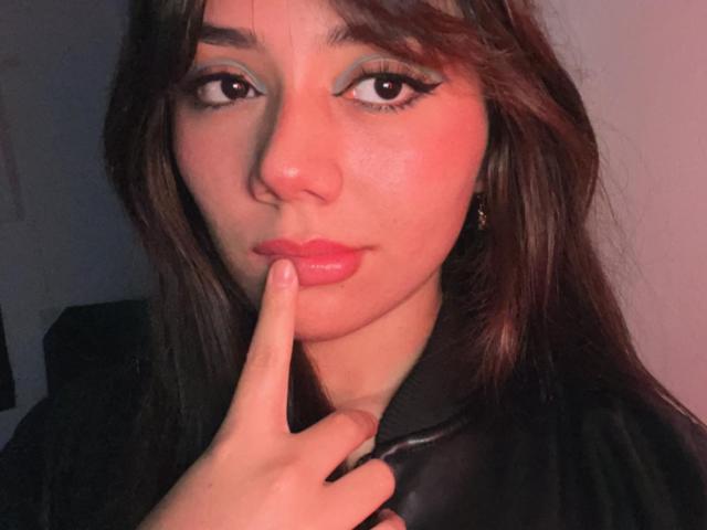 Alanna Jonson, com lábios rosados, dedo na boca, cabelo escuro, jaqueta de couro preta, delineador azul e brincos dourados, com uma aparência sedutora e rosto levemente corado..