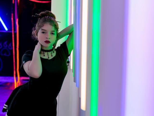 EvaRavissante leans against a neon-lit wall, wearing a black dress with a white, beaded choker, her hand on her neck, green and blue lights highlighting her curvy ass.