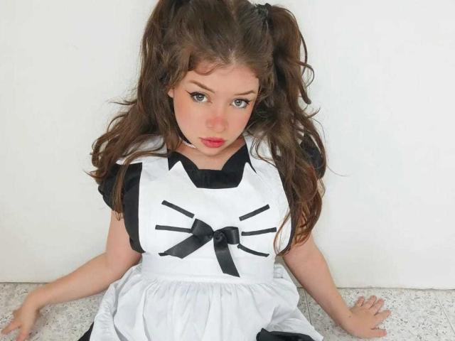 EvaRavissante with long brown pigtails, wearing a black and white maid outfit, sitting on a floor, hands down, looking up with red lips and eyeliner.