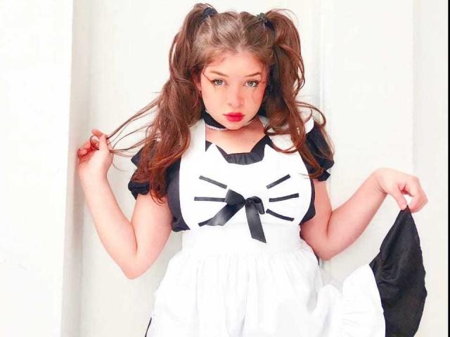 EvaRavissante with puffy brown twin-tails, black and white maid outfit, holding black panties, white wall background, red lipstick, black ribbon, and seductive gaze.