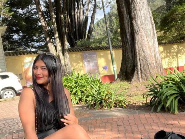 SexyDanyLove con cabello negro largo, lápiz labial rojo, top negro sin mangas y gafas en la cabeza, sentada en un camino de ladrillos, sonriendo, rodeada de árboles y plantas verdes, un día soleado..