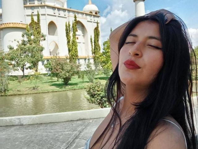 SexyDanyLove com cabelo preto, olhos fechados, batom vermelho e a mão no cabelo, vestindo uma blusa cinza, em pé perto de um lago, com um prédio branco com cúpula ao fundo..