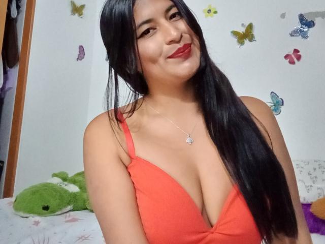 SexyDanyLove with long black hair, red lipstick, and a low-cut orange top, showing her big tits and smiling at the camera. Butterfly stickers on the wall.
