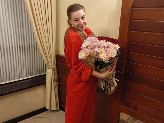 SweetyMollyyss in een rode jurk, met een boeket roze en witte bloemen, lachend met haar blonde haar in een staart, staand in de buurt van een houten deur..