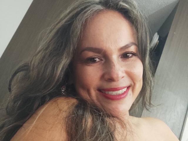 Danny69s tem cabelos grisalhos longos e ondulados, olhos castanhos e um sorriso vermelho que mostra seus dentes. Ela está sorrindo para a câmera com os ombros descobertos. Seus seios estão parcialmente visíveis..