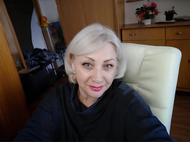 Tashyncik with short white blonde hair, blue eyes, pink lipstick, wearing a black shirt, sitting on a white chair, smiling at the camera, showing her face and upper body.