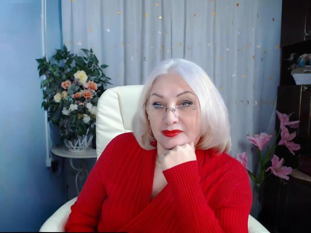 Tashyncik with pale blonde hair, red lipstick, glasses, and red sweater, resting chin on hand, looking at camera. She has medium-sized tits, sitting in white chair.