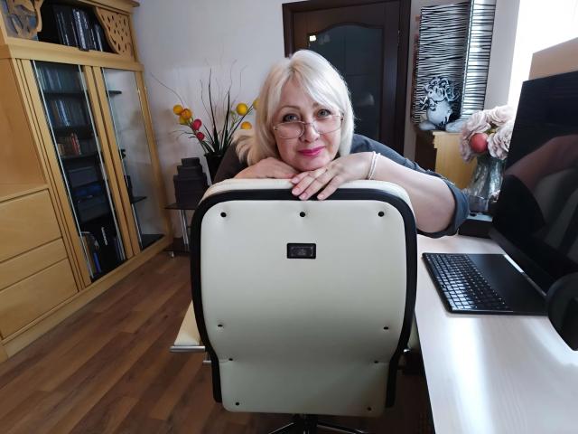 Tashyncik with blonde hair and glasses, leaning on a white office chair, showing her tits, in a wooden-floored office with a computer and yellow flowers.