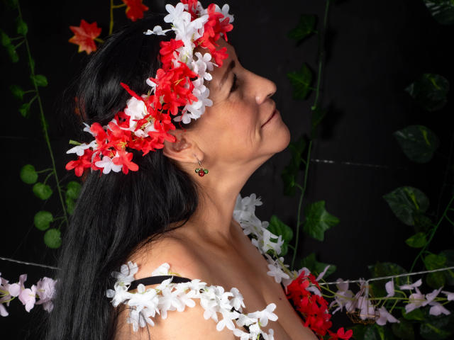 ScarlettSweet with black hair, wearing a red and white flower headpiece, and matching flowers on her shoulders, has her tits slightly visible, and a nose ring, facing right.
