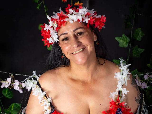 ScarlettSweet, a curvy woman with tan skin, black hair, and red/white flower crown, smiling, showing her boobs, adorned with floral straps, wearing green earrings.