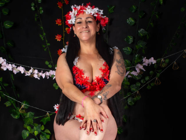 ScarlettSweet with red and white flower headpiece, red flower bra, black long hair, smiling, tattooed arms, pink panties, red nails, black background, green vines, and white flowers.