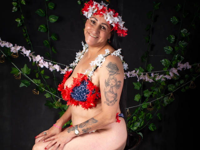 ScarlettSweet, middle-aged woman with tattoos, smiling, wearing red and white flower headpiece, blue and red feathered bra, and matching panties, sitting, black background, leafy green vines.