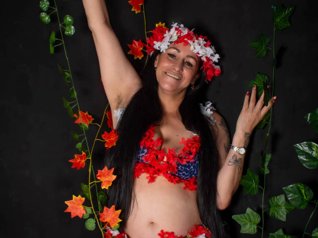 ScarlettSweet with long black hair, red and white flower headpiece, red flower bra, arm raised, smiling, tattoos on left wrist, green leafy vines, black background, watch on right wrist.