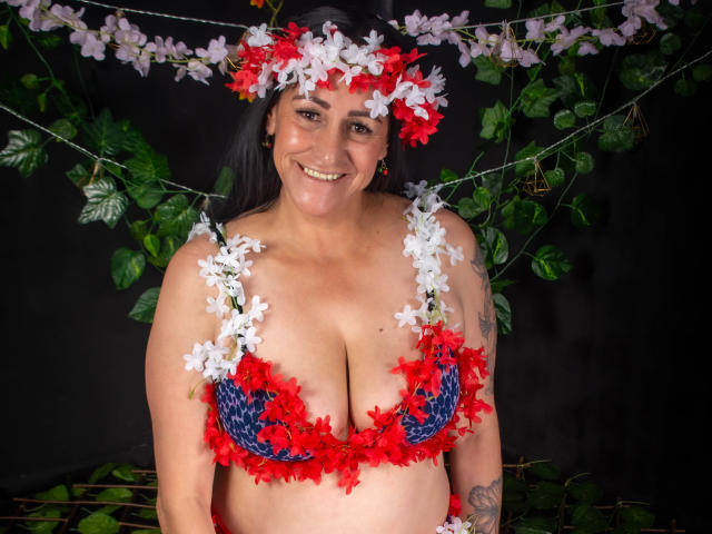 ScarlettSweet has large tits, wearing a red and blue floral bra, with red and white flowers in her hair, smiling, tattoo on right arm, dark hair, black background.