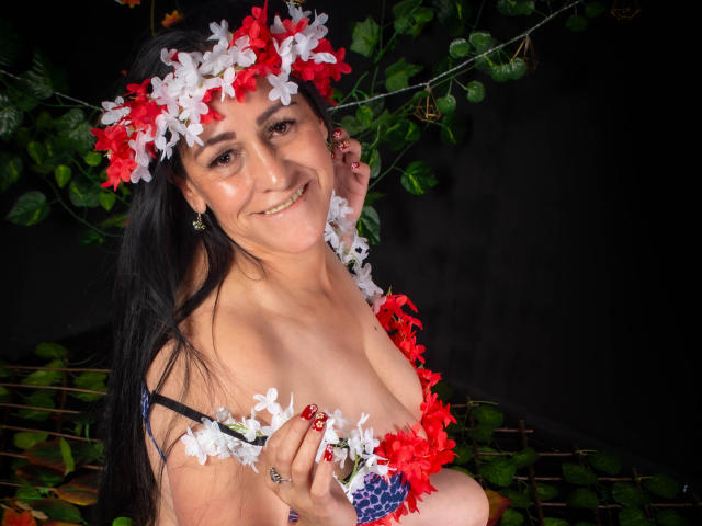 ScarlettSweet with long black hair, wearing a floral headband and bra, smiling at the camera, holding flowers over her tits. Dark background, green leaves, and a metal railing.