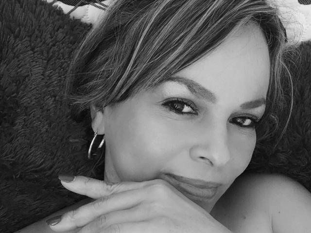 Black-and-white photo of Danny69s with short, light-colored hair, hoop earrings, and subtle makeup, resting her chin on her hand, showing her lips and looking slightly to the side.