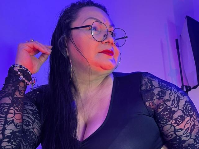 EllenaBoobs with glasses, black lace sleeves, and red lipstick, holding her wet black hair, wearing a black V-neck top, purple and blue lighting, and silver bracelets.