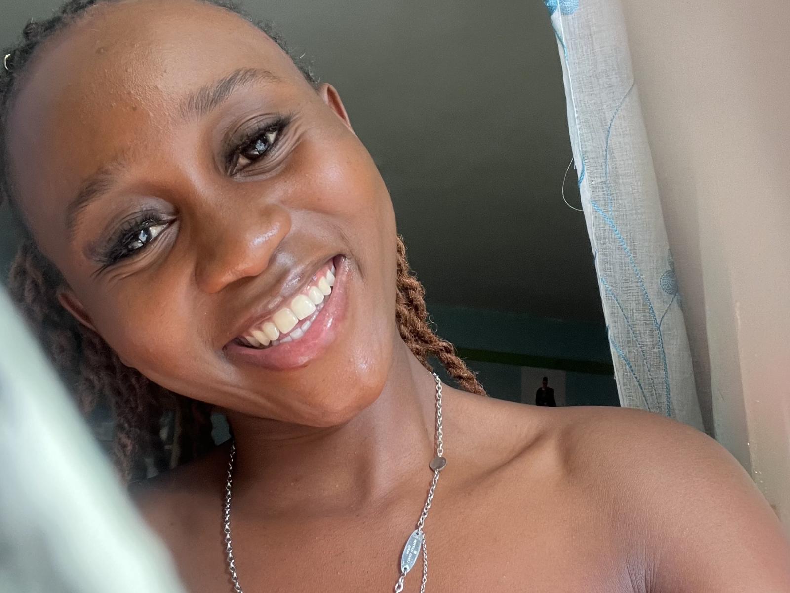 BarbaraFly with dark brown skin, smiling with teeth showing, wearing a silver necklace, has braided hair, and is topless, looking at the camera.
