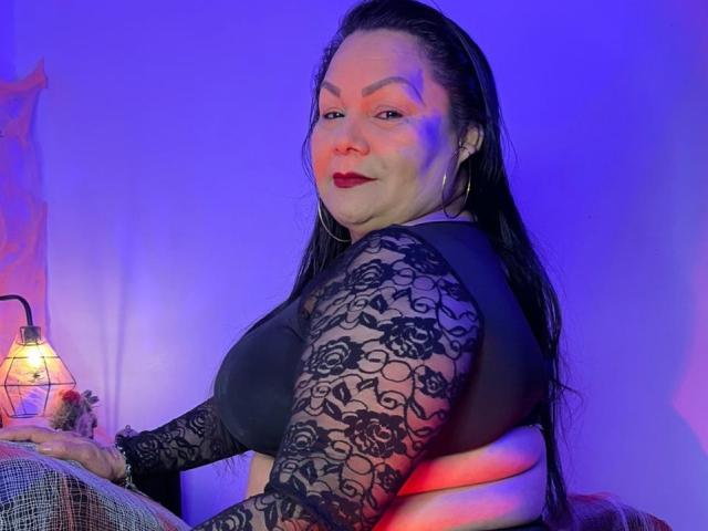 EllenaBoobs with long black hair, red lipstick, wearing a black lace top, showing her tits, sitting with a lamp and patterned pillow, under blue and purple lighting.