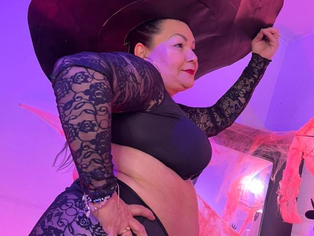 EllenaBoobs with red lipstick, black lace top, and matching panties, holding a large black hat, showing her curvy ass, in a purple-lit room, wearing a bracelet.