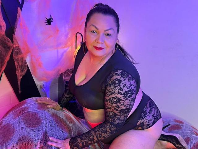 EllenaBoobs with dark hair in a ponytail, wearing black lace lingerie, sitting on a bed with pink and purple lighting, showing her ass and tits, smiling at the camera.