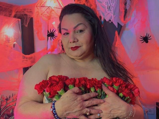 EllenaBoobs with red roses, holding them against her tits, has red lipstick and long dark hair, wearing bracelets, in a colorful, spooky room with orange and pink lights.