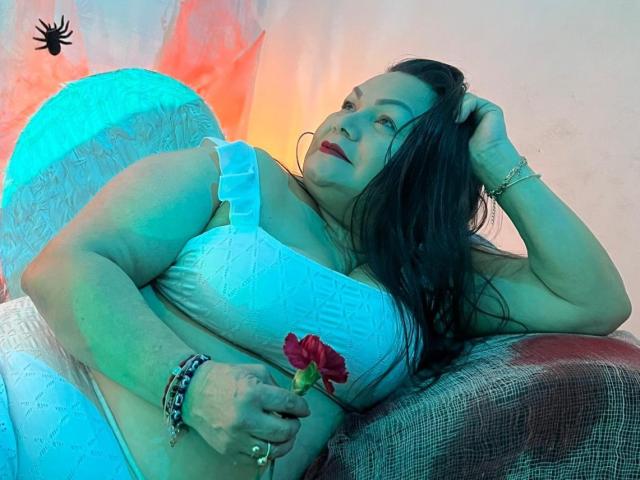 EllenaBoobs has big tits, wearing a white lace bra, lying on a red and brown textured surface, holding a red rose, with blue and orange lighting, and dark hair.