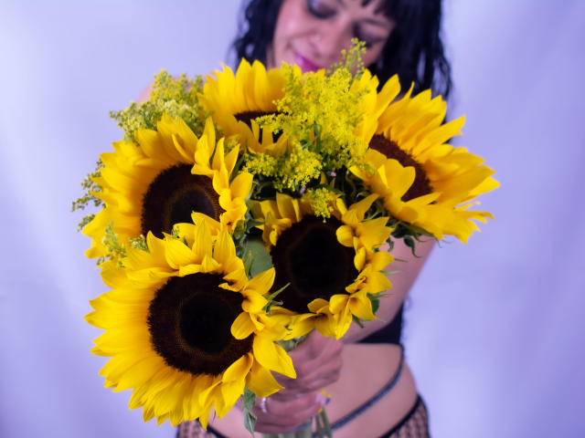  AlexandraJones holding a bouquet of bright yellow sunflowers with dark centers and green yellow foliage, smiling slightly, wearing black lingerie, in a colorful, vibrant photograph.