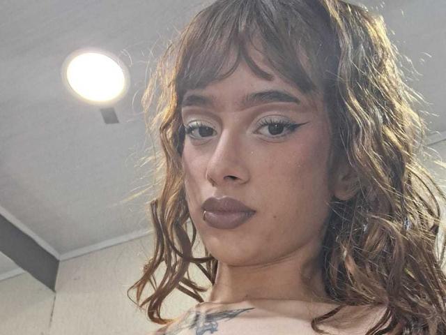 CamiMonet with wavy brown hair, dark lipstick, and eyeliner, has a nose ring and tattoo on her chest, looking at the camera in a selfie.