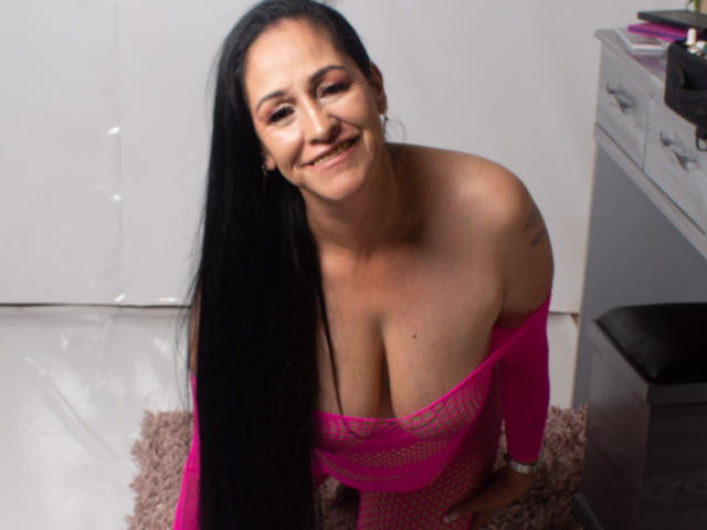 ScarlettSweet with long black hair, smiling, wearing a pink mesh off-shoulder top, exposing her large tits, sitting on a pink rug in a room.
