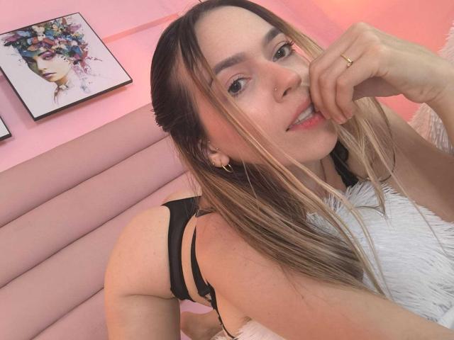 KatieSidney with blonde hair, black strappy lingerie, white furry blanket, gold hoop earrings, nose ring, smiling, hand on chin, pink bedroom, framed art, ring on finger, cute.
