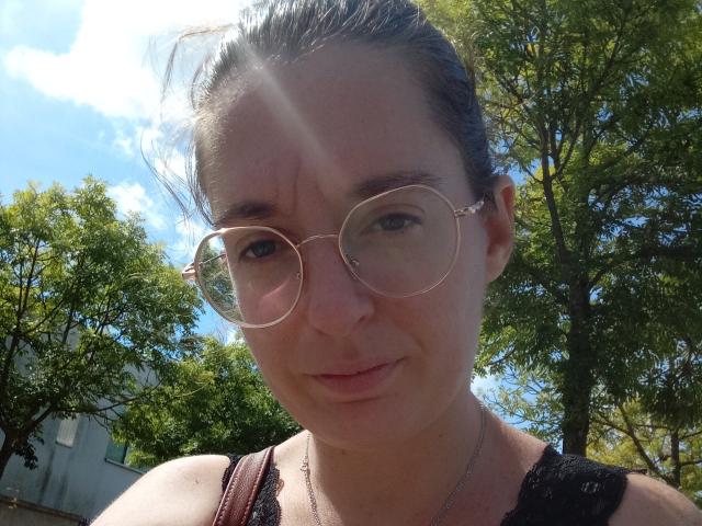 JolieJoelle with small round glasses, light skin, brown hair in a bun, black sleeveless top, subtle smile, sunbeam on face, necklace, and green trees in the background.