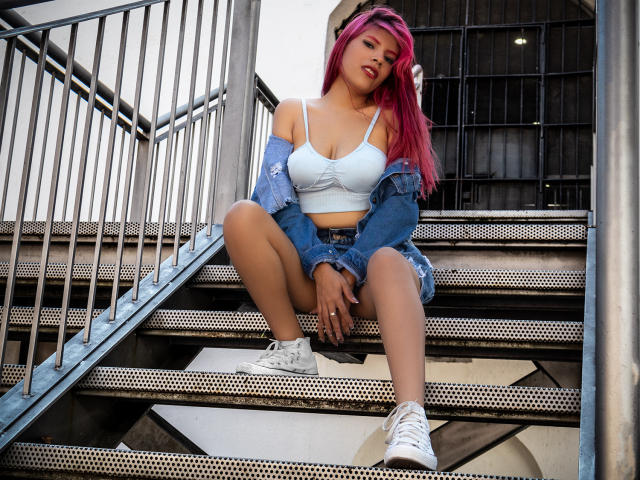 AfroditaMooreX with pink hair, white bra, denim jacket, and shorts, squatting on metal stairs, white sneakers, exposing her tits and ass, looking down with a relaxed expression.