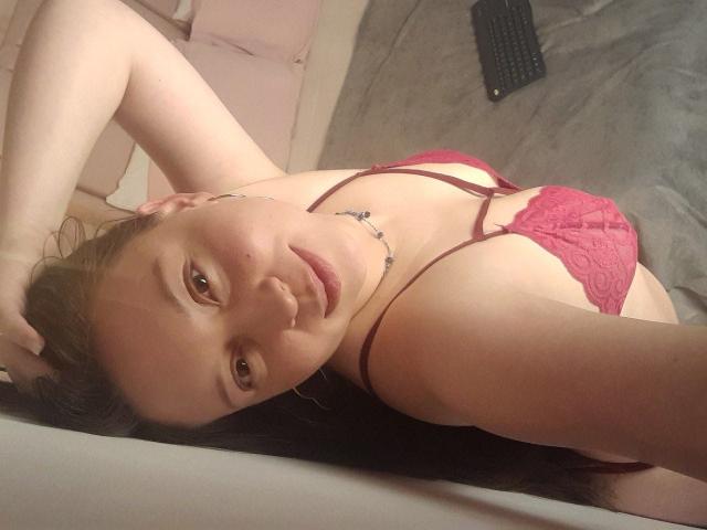 WidnyBeaumont ligt op een bed, ze draagt een rode kanten beha, met haar arm omhoog, waarbij haar borsten, bruine ogen en een tatoeage onder haar kin zichtbaar zijn..