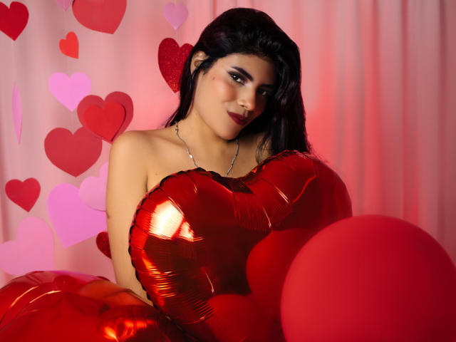 AfroditaMooreX with shiny red heart-shaped balloons, holding a large red heart, wearing a strapless red outfit, black hair, red eye makeup, and a subtle smile.