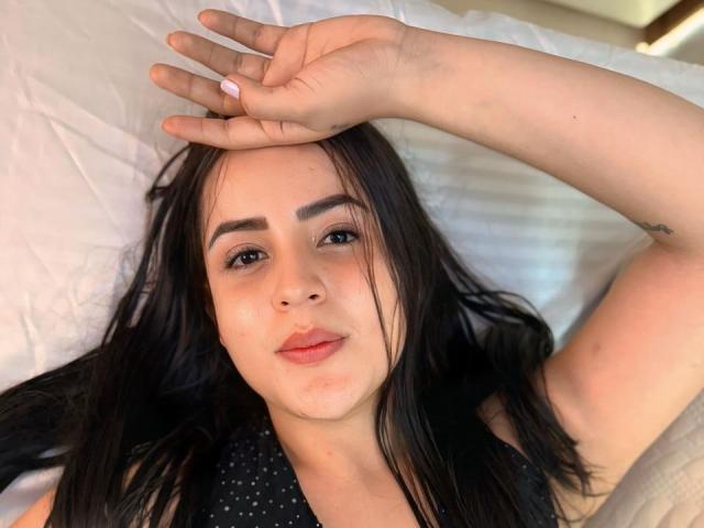 DannaMorel tem cabelo escuro, olhos castanhos e uma expressão relaxada, deitada em uma cama com o braço sobre a cabeça. Ela está vestindo uma blusa preta com bolinhas..