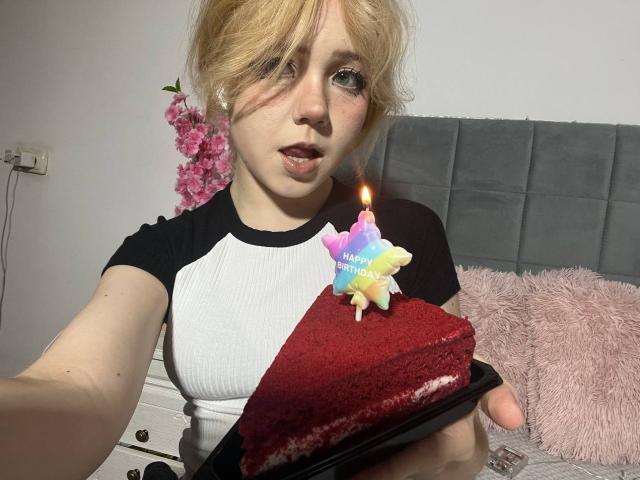 EmilyAngels with messy blonde hair, wearing a black and white raglan shirt, holding a red slice cake with a candle, looking at the camera, in a bedroom.