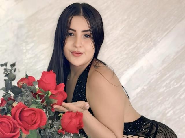 DannaMorel mit langen, schwarzen Haaren, die ein schwarzes, glitzerndes Top mit Halstuch trägt, rote Rosen hält, lächelt, hat helle Haut und ein kleines Tattoo an ihrer rechten Schulter..