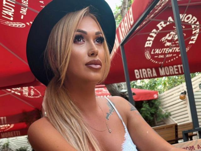 NicolSNaya with blonde p Ponytail, black hat, and white lace top, showing a small tattoo on her right shoulder, looking off-camera with a subtle smile.