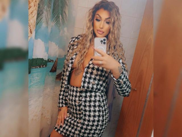 NicolSNaya with long curly hair, wearing a black-and-white houndstooth dress, showing cleavage, holding a phone, mirror selfie, tattoos visible on chest. Boobs, dress, phone, tattoos.
