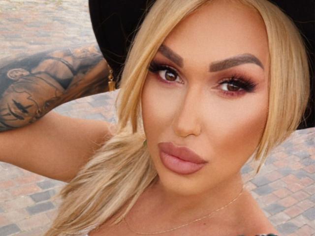 NicolSNaya has blonde hair, dark eyebrows, red eyes, and pink lips. She's wearing a black hat, showing a tattooed arm. Boobs are partially visible.