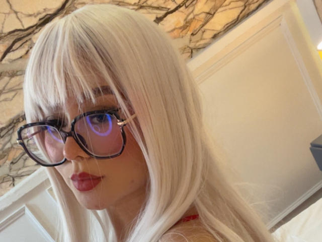 Millie Reed, avec ses cheveux blancs, ses lunettes noires et ses lèvres rouges, montrant son visage et une petite partie de sa poitrine, dans un autoportrait avec des lunettes réfléchissantes..