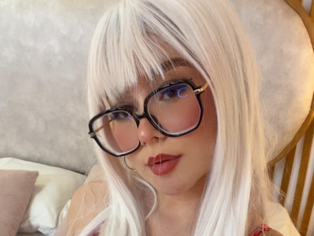 Millie Reed mit weißem Haar, schwarzen Brillen und pinkem Lippenstift, die ihre Brüste und ihr süßes Gesicht zur Schau stellt. Sie schaut direkt in die Kamera mit leicht geöffnetem Mund..